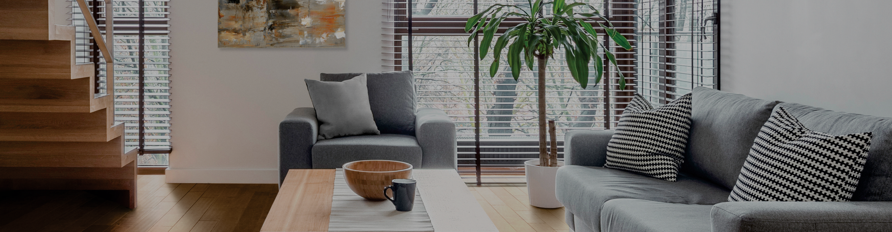 A modern living room features a wooden staircase, a grey sofa with striped pillows, a grey armchair, a coffee table with a bowl and mug, and a potted plant near a window with blinds.